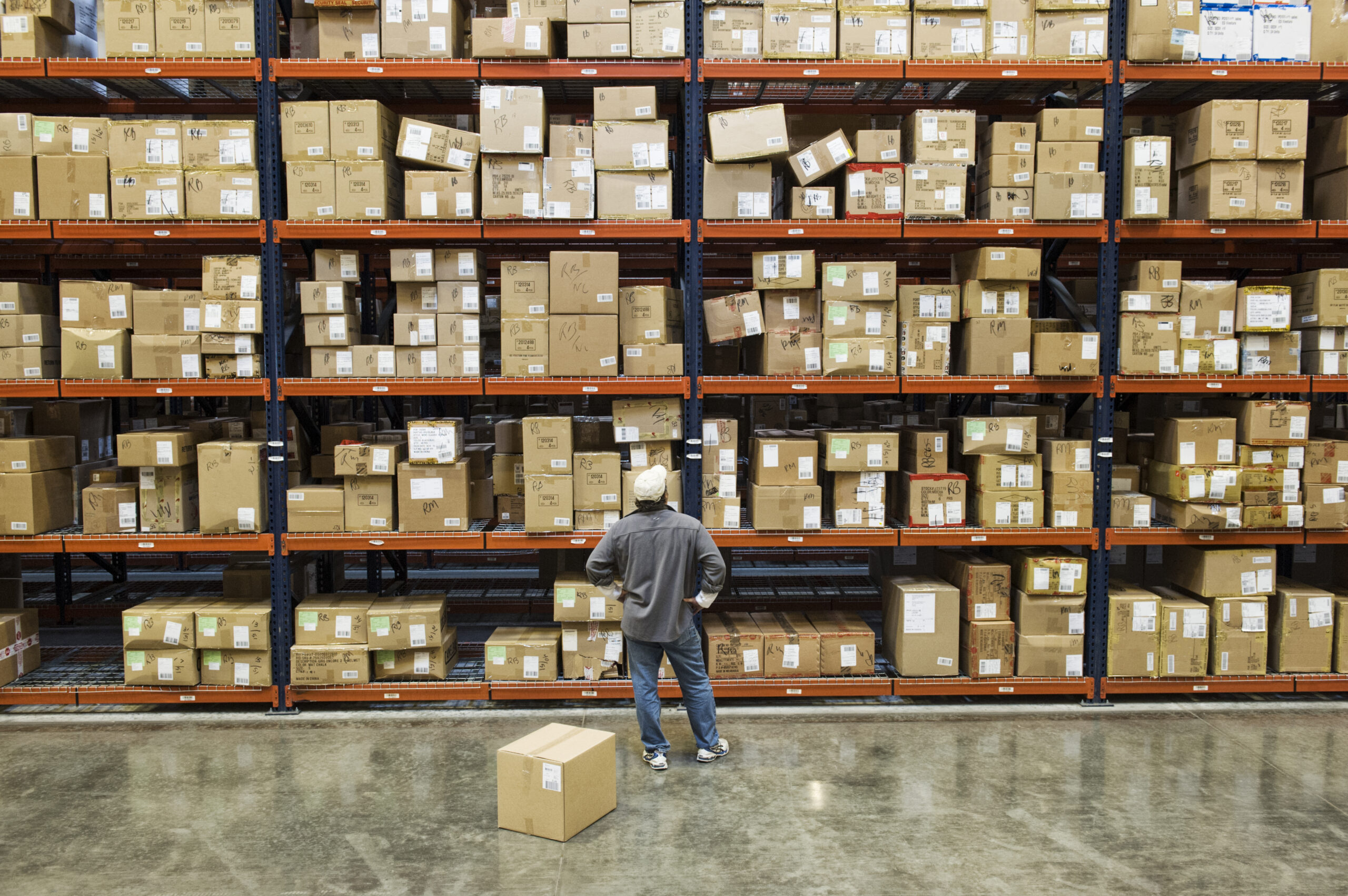 Introduction to Lean Warehouse worker checking inventory next to large racks of cardboard boxes holding product in a distribution warehouse.